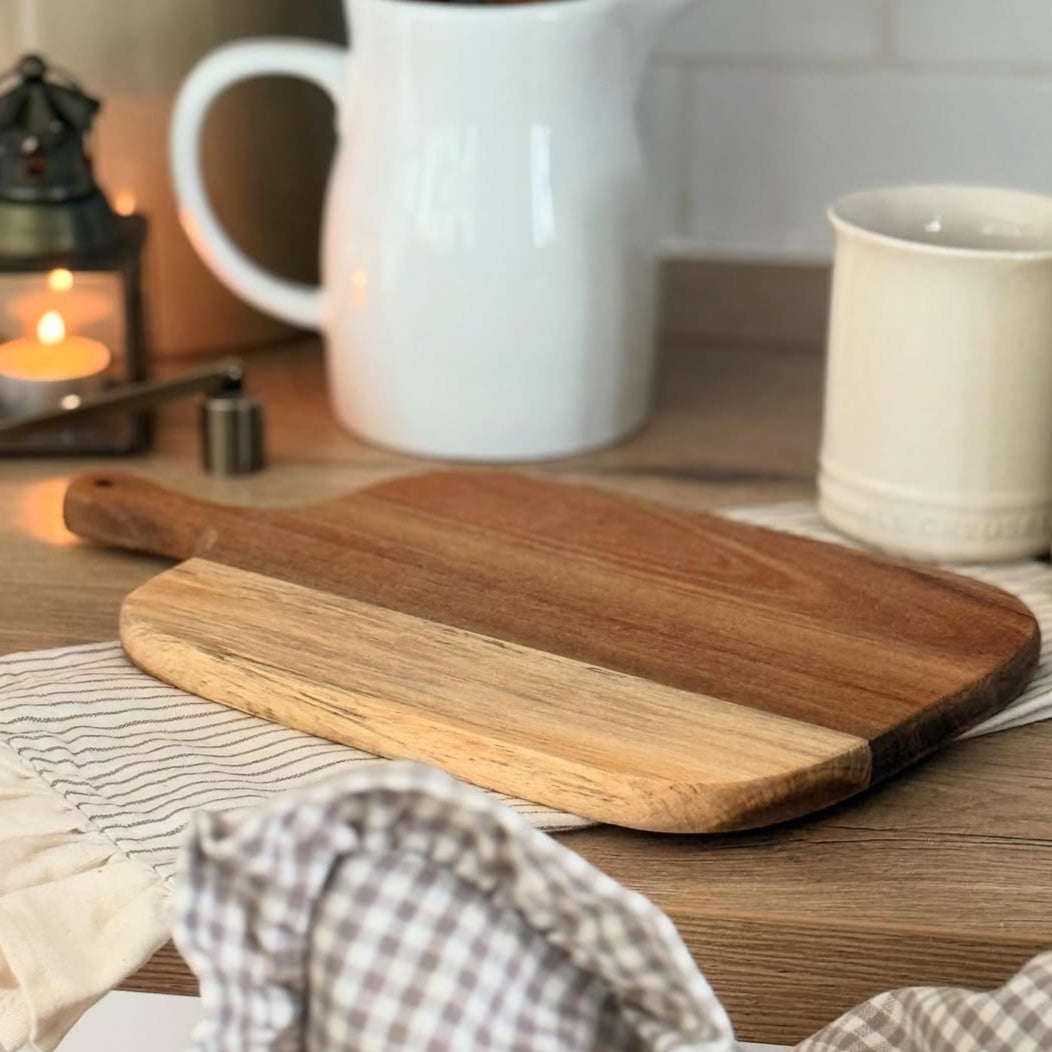 Wooden acacia cutting board on a kitchen counter with a checkered cloth and ceramic items.