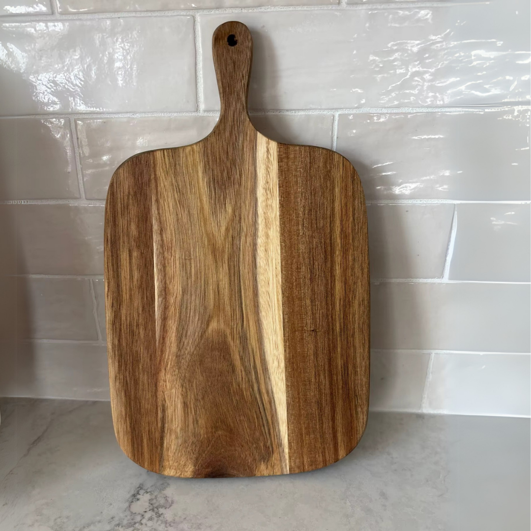 A rectangular wooden chopping board with a handle, placed against a tiled wall on a marble surface.