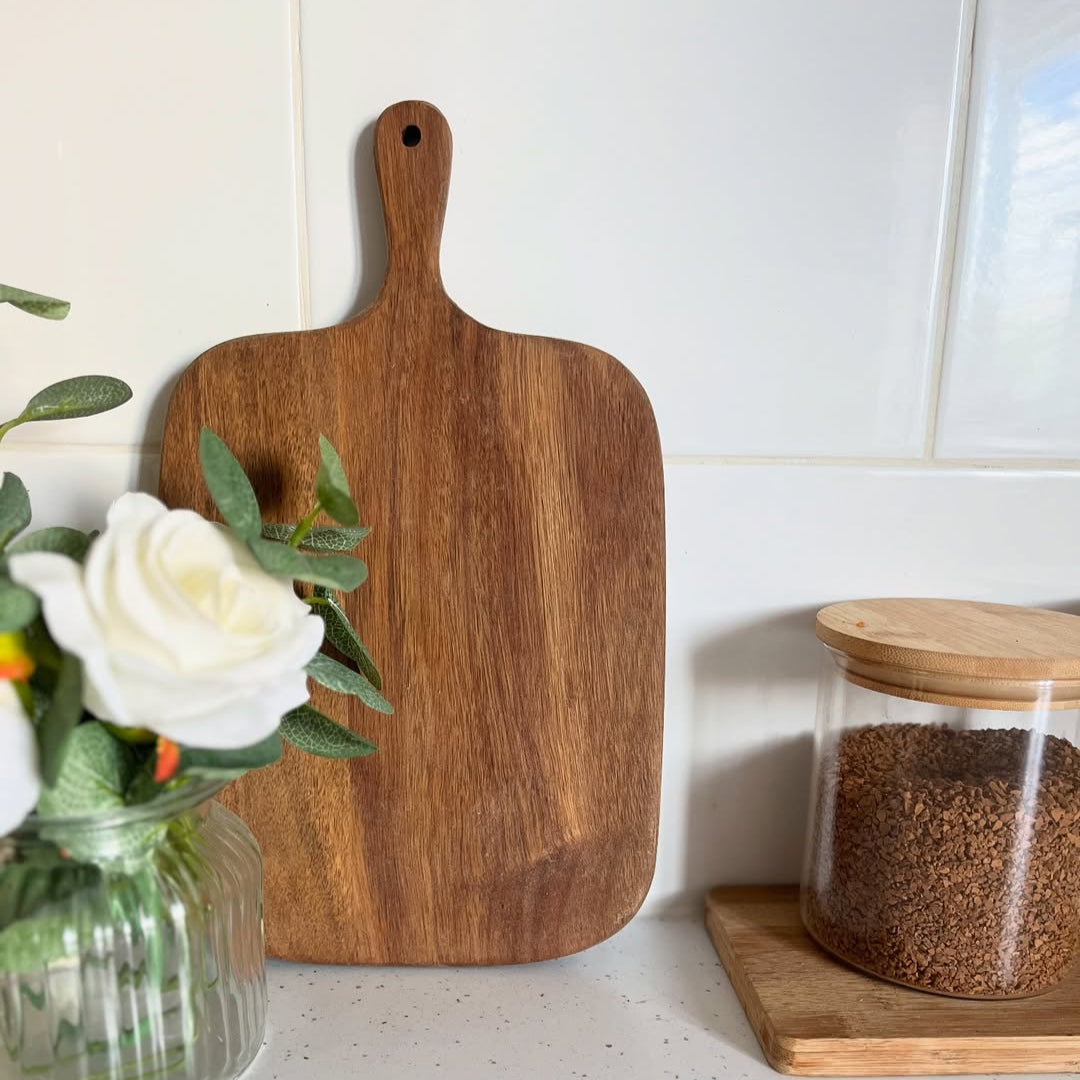 Wooden cutting board on a kitchen counter with a vase of flowers and a jar.