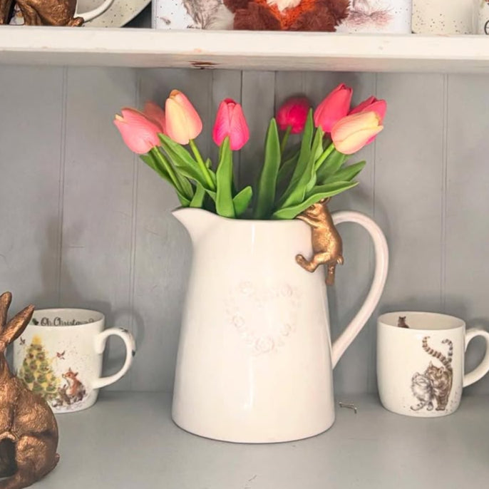 White pitcher with pink tulips on a shelf with decorative items