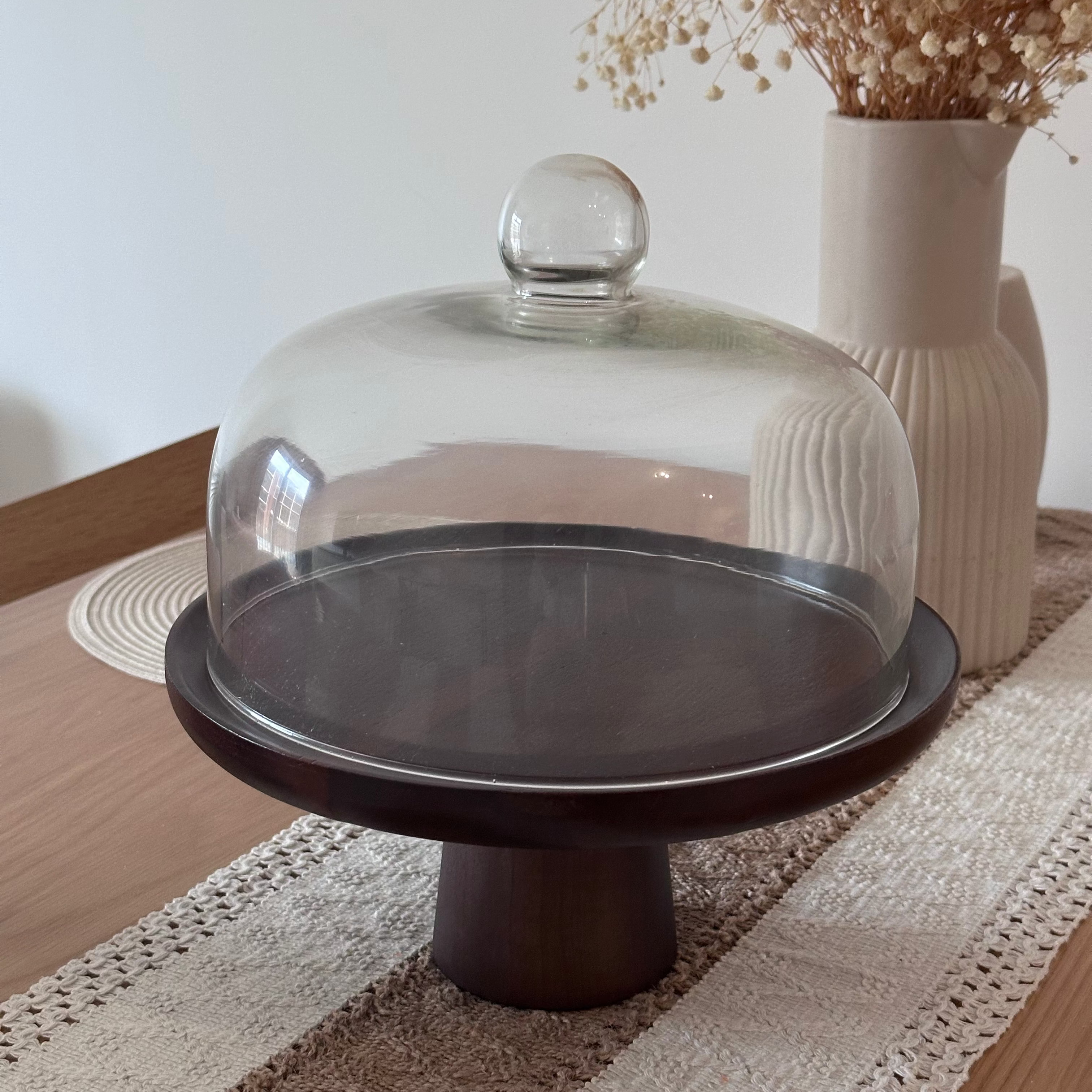 Decorative cake stand with glass dome on a table with a vase in the background