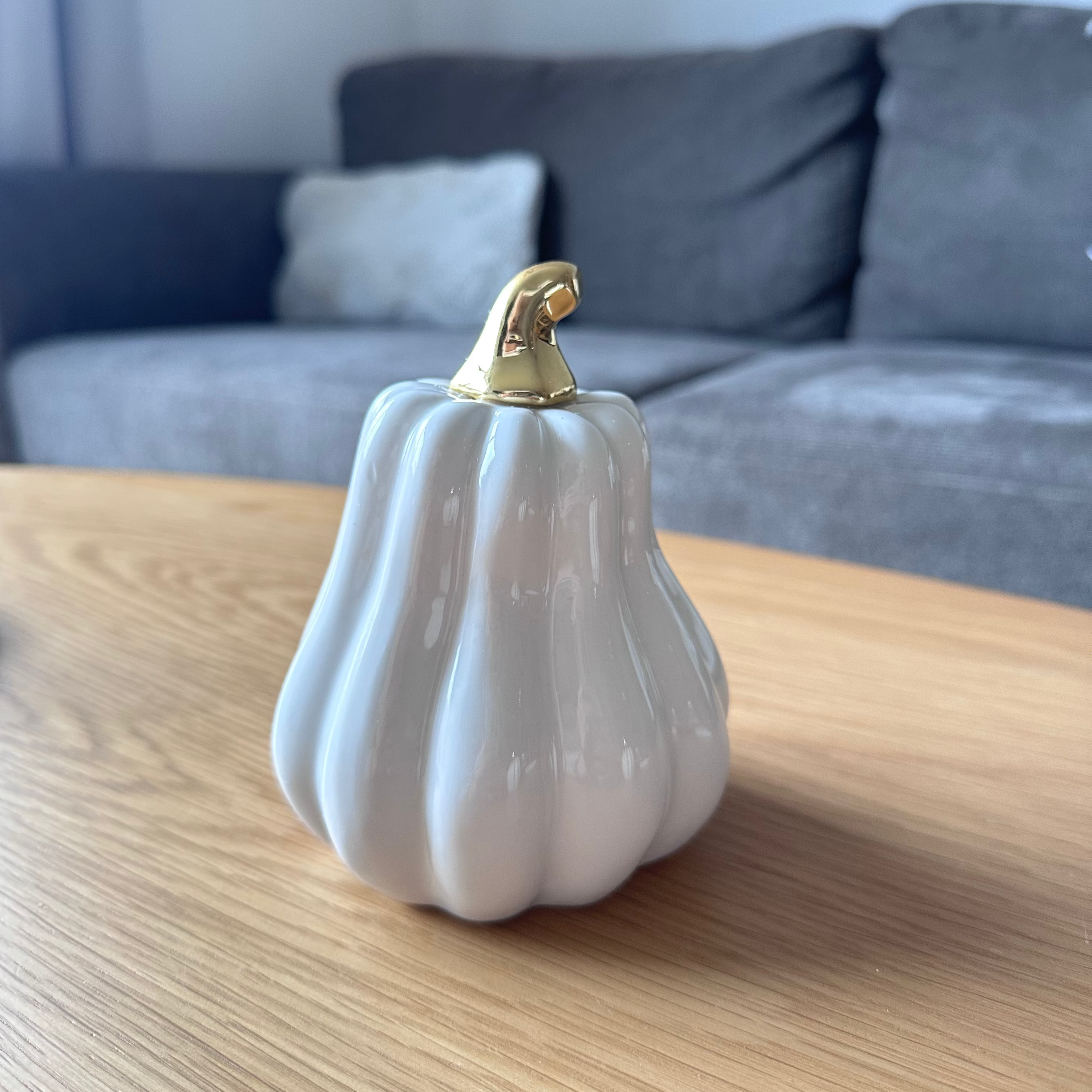 White ceramic pumpkin with gold top on a wooden table, with a gray sofa in the background.