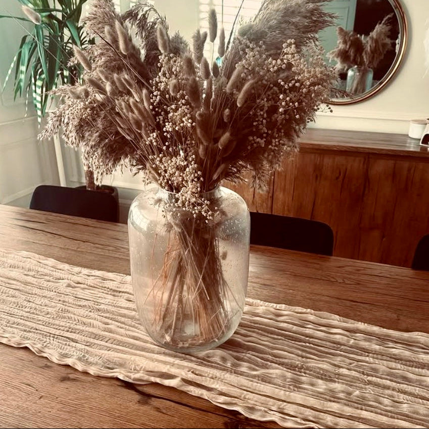 Clear vase with dried plants on a wooden table, where a cheesecloth rustic table runner is featured.