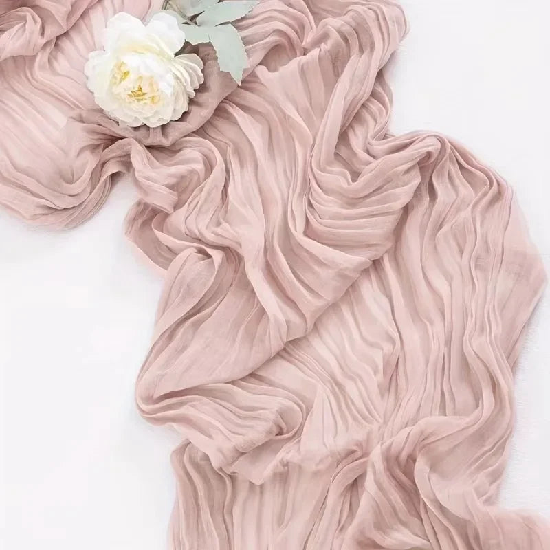 A close-up of a light pink cheesecloth rustic table runner draped on a white surface. A single flower also features.