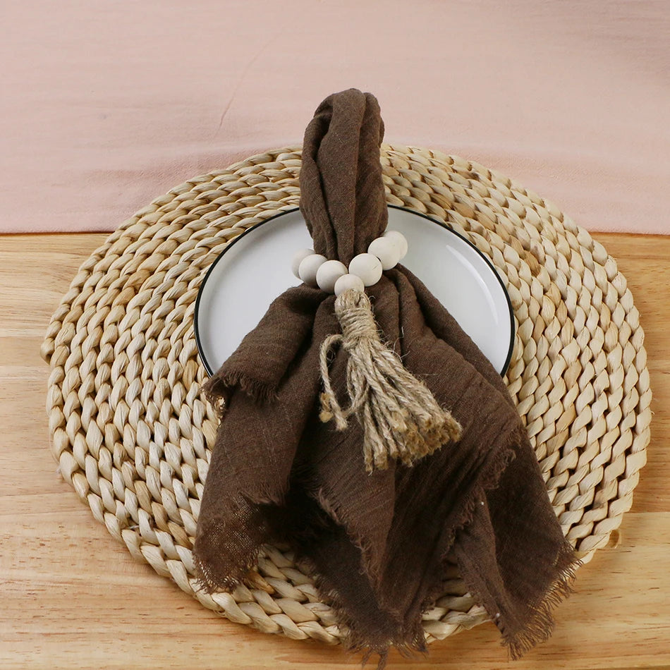 A brown cotton napkin is draped over a woven round table, with a white plate and a decorative bead on the napkin, set against a pink background.