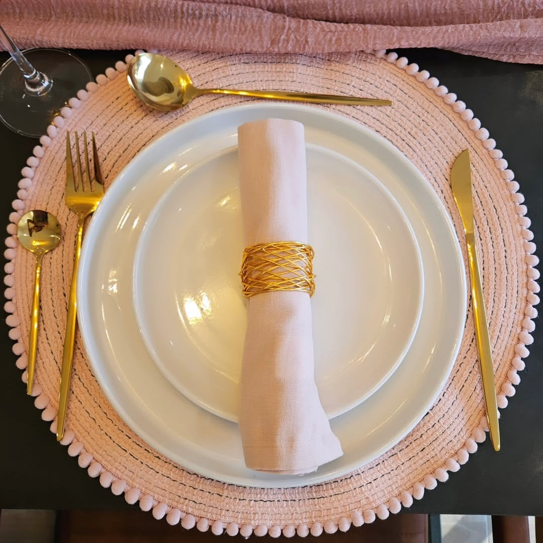 Dinner setting with white plates, gold cutlery, and a pink napkin on a woven placemat.
