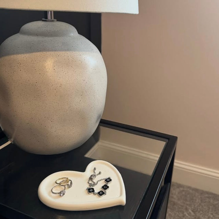 Heart-shaped jewelry dish with rings and earrings on a nightstand next to a lamp.