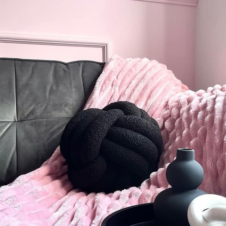 Black knot pillow on a pink textured blanket with a grey headboard.