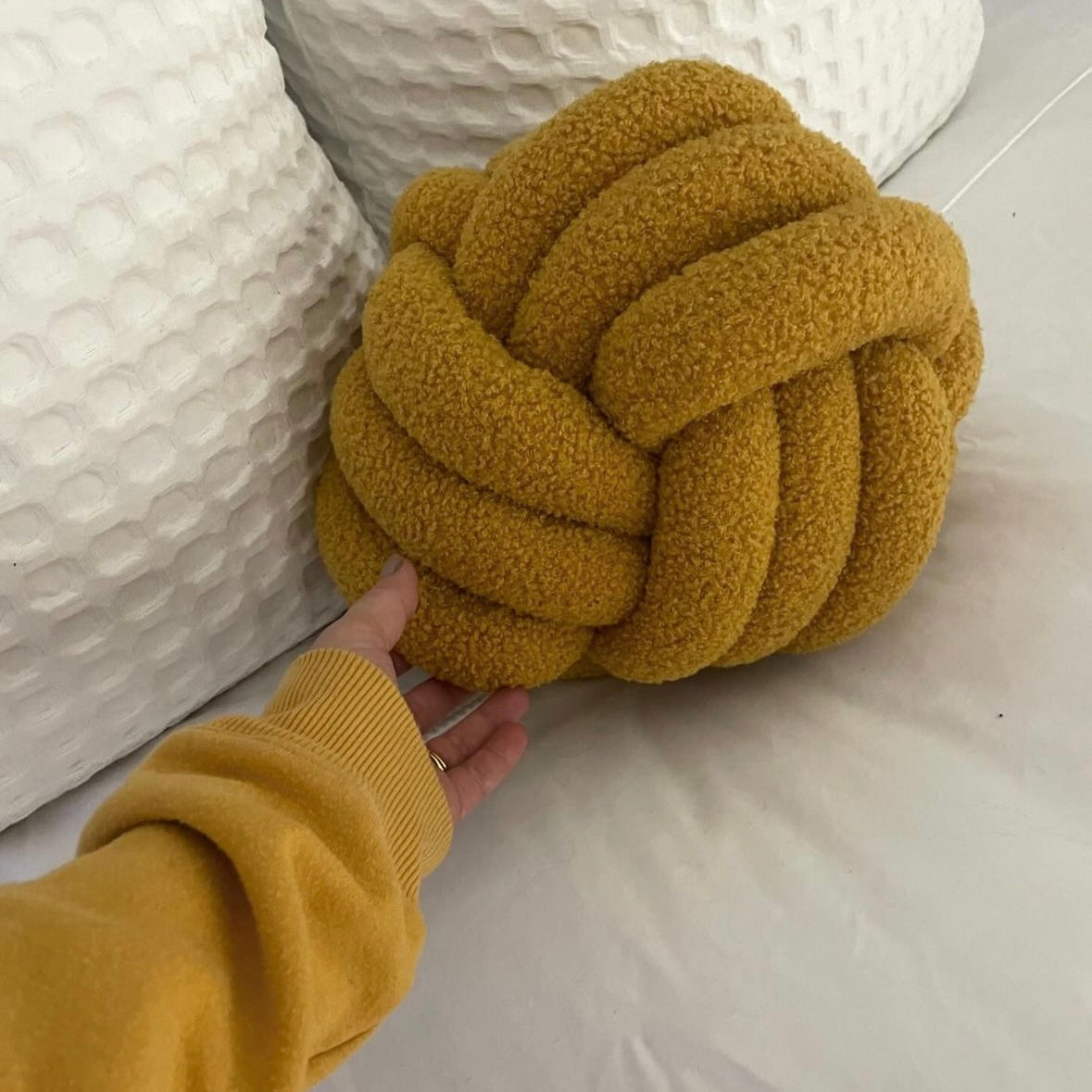 Mustard yellow knotted pillow held by a hand against a white textured background