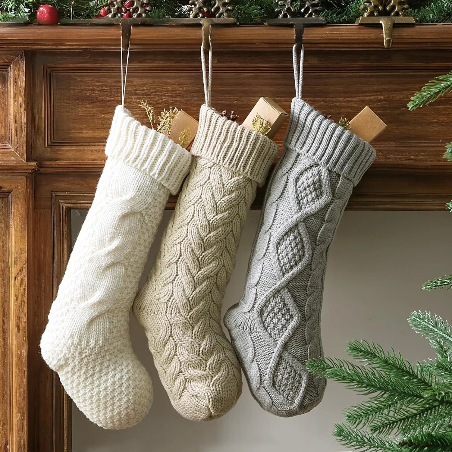 Three white, beige and grey cable-knitted Christmas stockings hanging from a wooden mantel, each with a festive design and a Christmas tree in the background.
