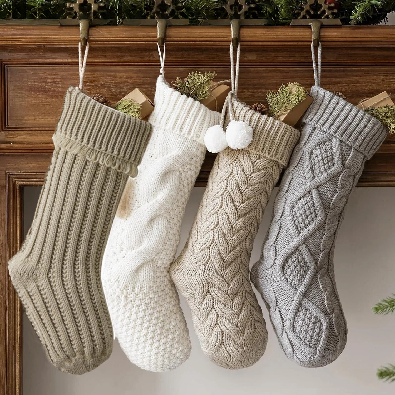 Four white, beige and grey cable-knitted Christmas stockings hanging from a wooden mantel, each with a festive design.