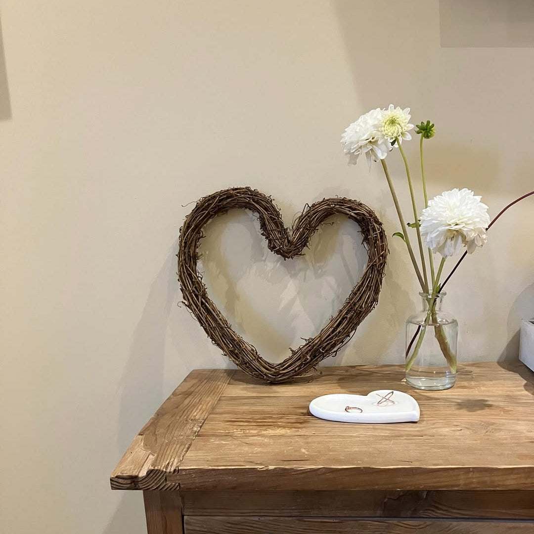Wooden table with a heart-shaped wreath and vase of flowers against a plain wall.