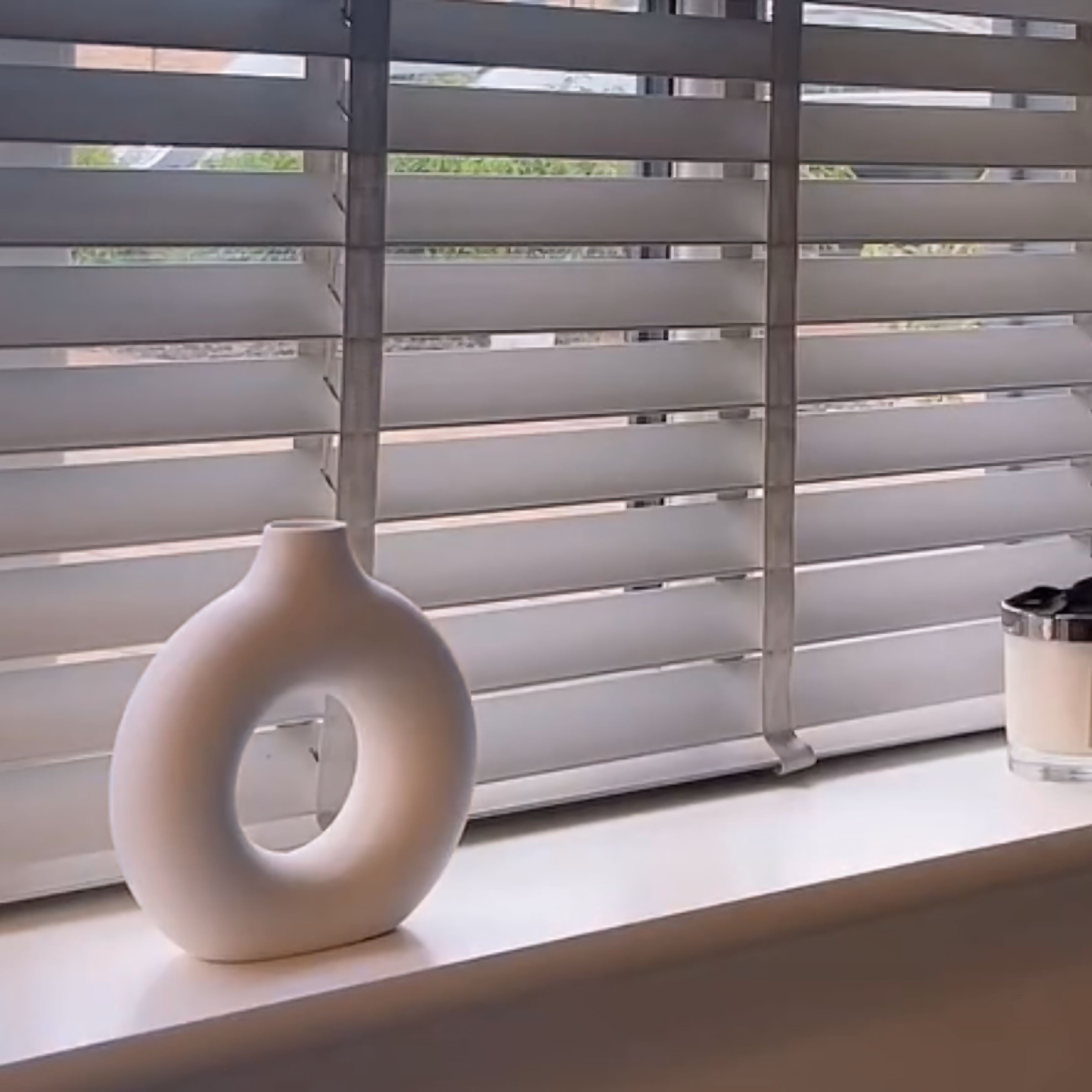 White nordic, circular vase on a windowsill with blinds in the background