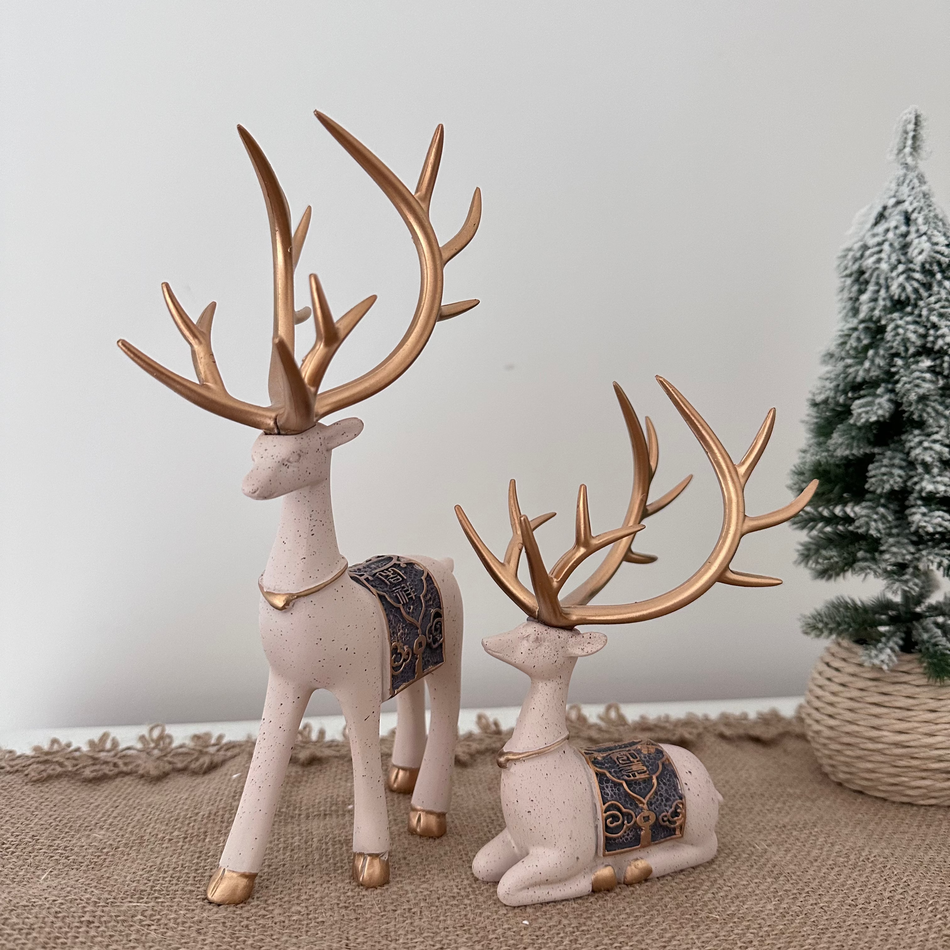 Two decorative deer figurines with gold antlers on a textured surface with a Christmas tree in the background.