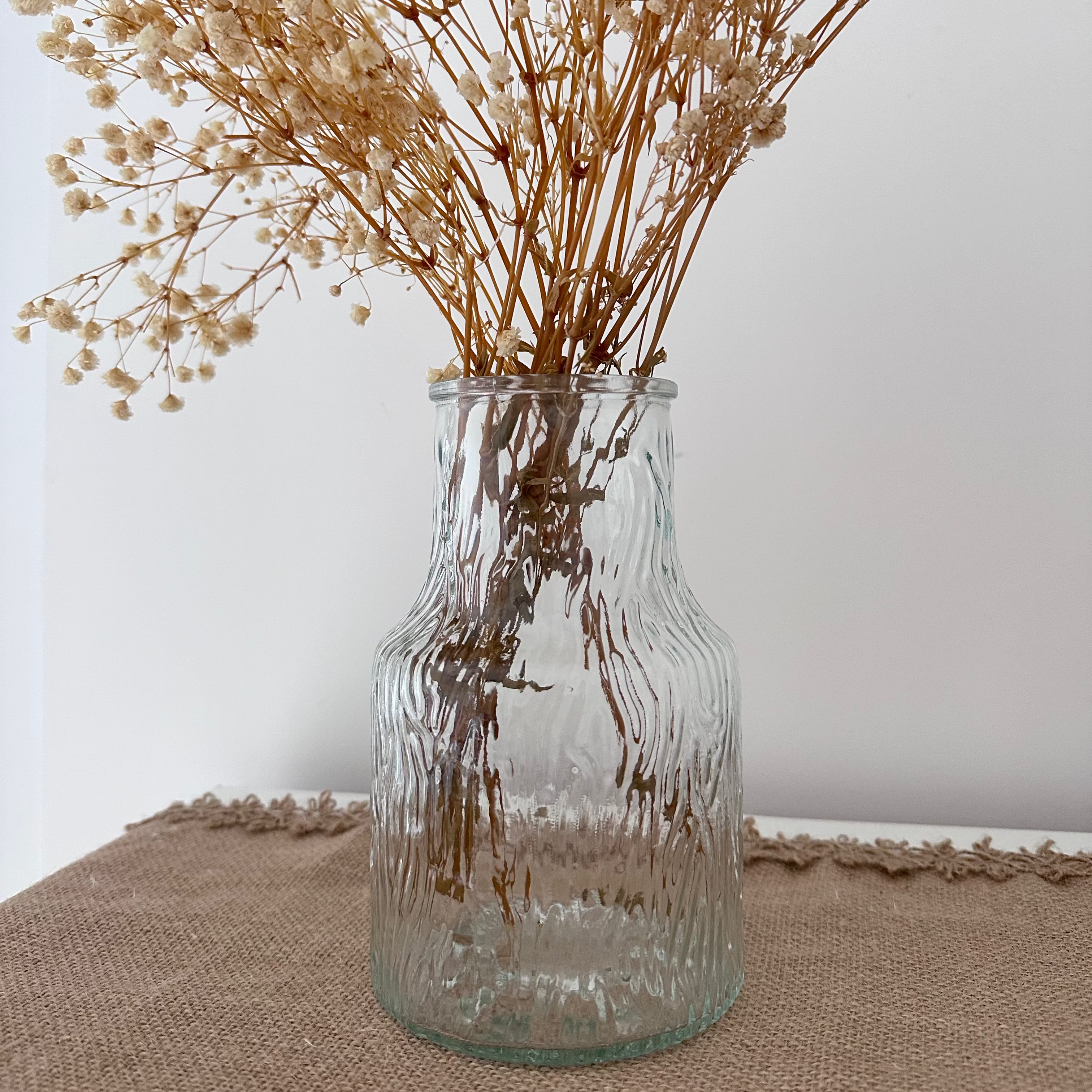 Clear, textured glass bottle with dried flowers on a hessian table runner
