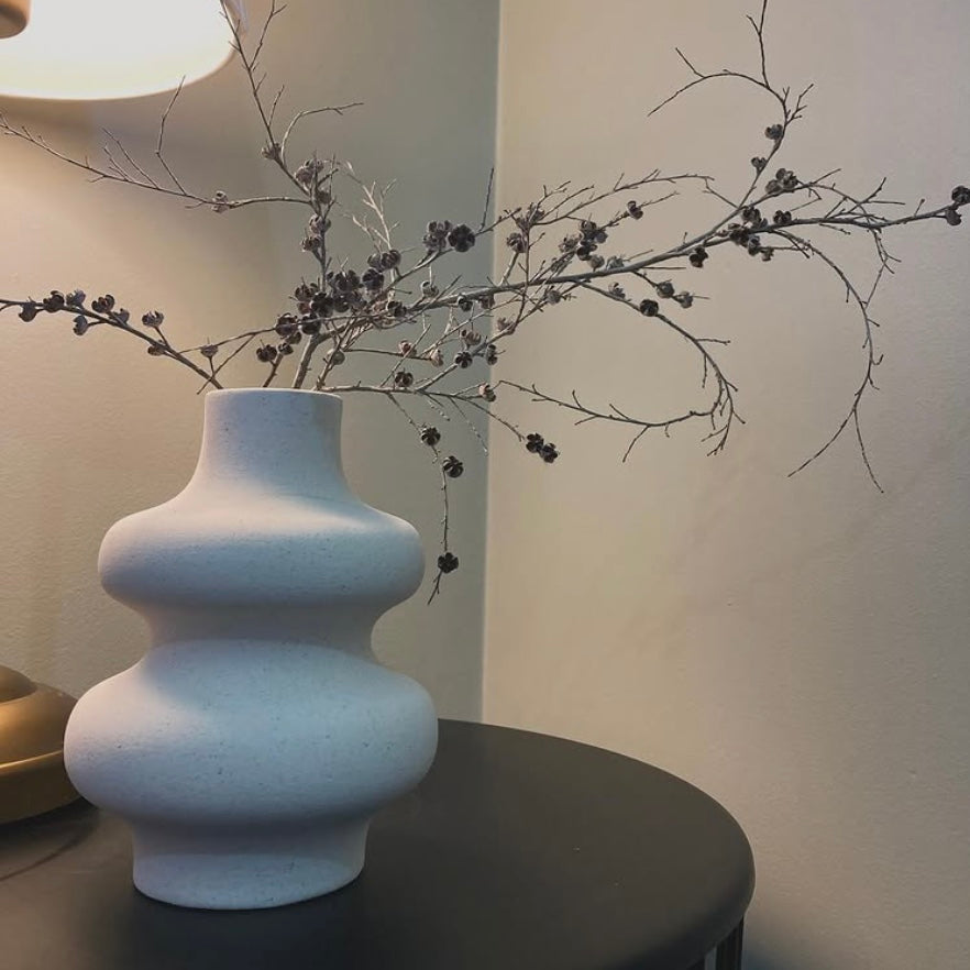 White, wavy ceramic vase with decorative branches on a dark surface against a neutral wall