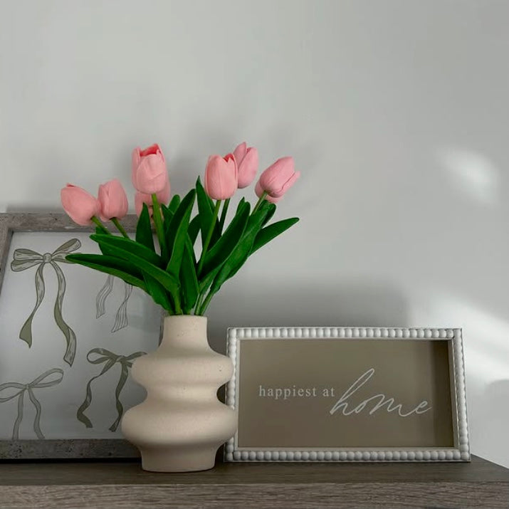 Pink tulips in a wavy, white ceramic vase on a shelf with decorative frames in the background.