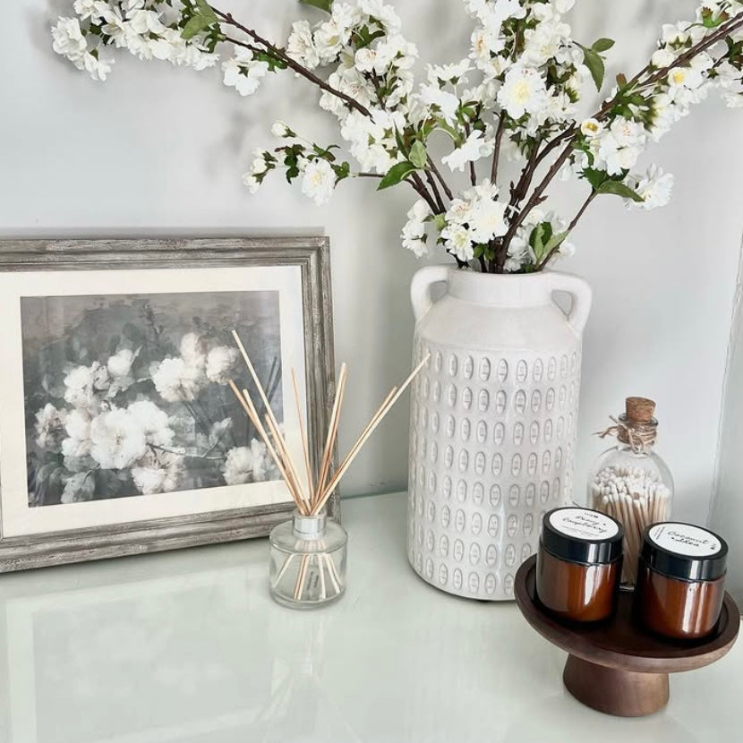 Decorative setup with a vase of white flowers, candles, and a diffuser on a shelf. A small wooden cake stand has been repurposed to support scented decorative candles.