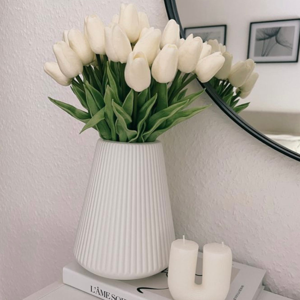 White tulips in a white vase on a surface with a mirror and books in the background.