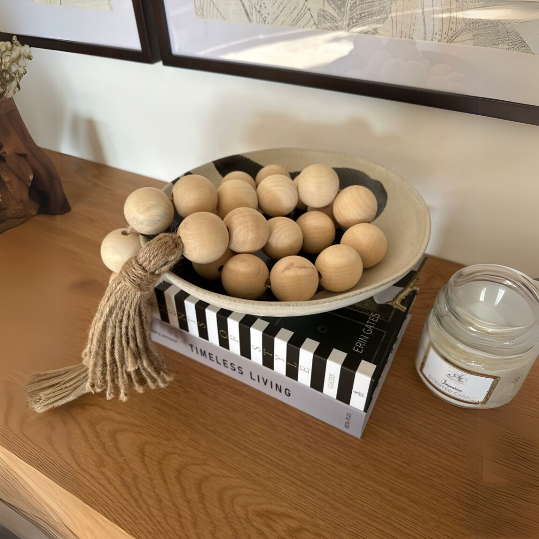 Decorative bead garland with hemp tassel styled in a wooden bowl on a table next to a candle and books.