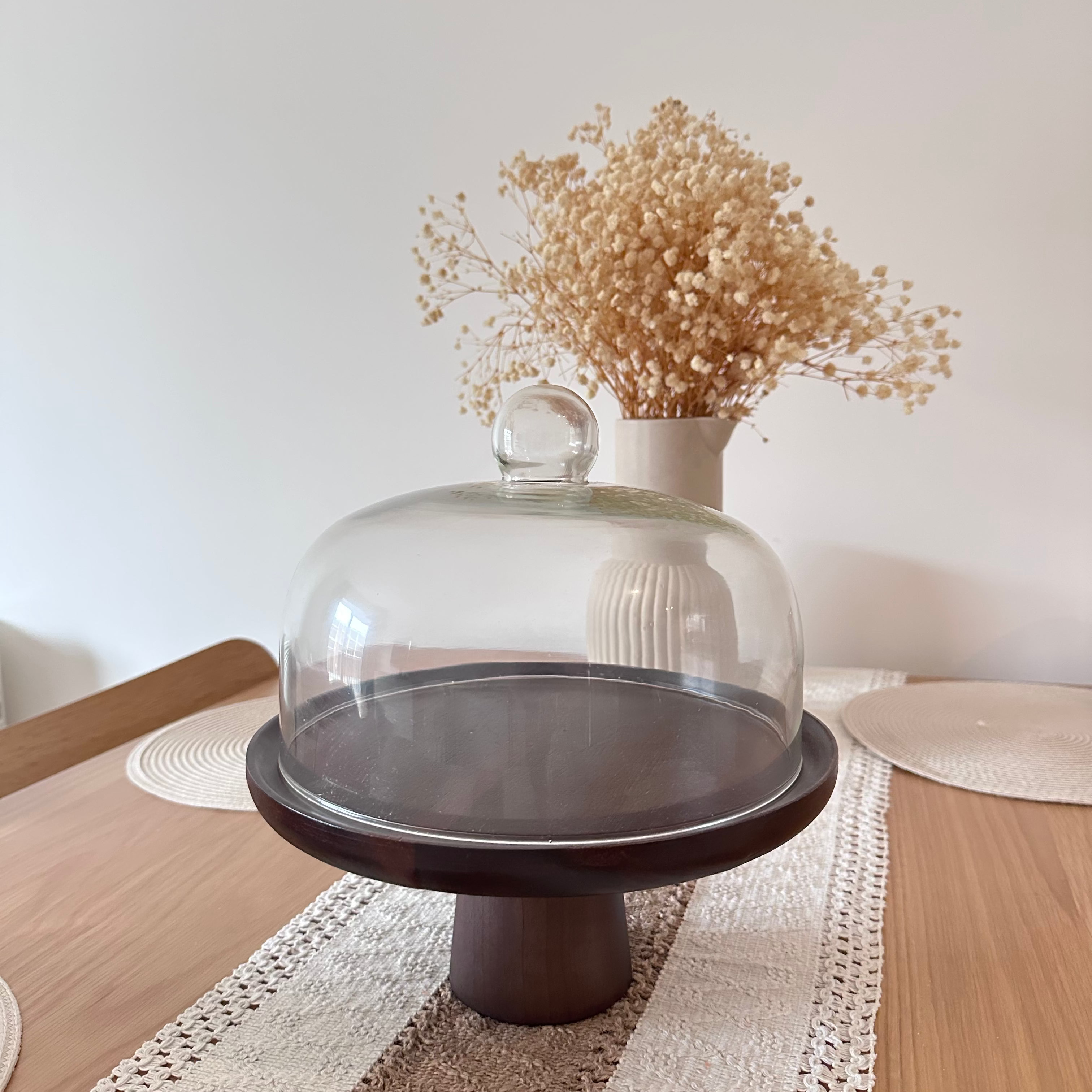 Decorative cake stand with a glass dome and vase of dried flowers on a table.