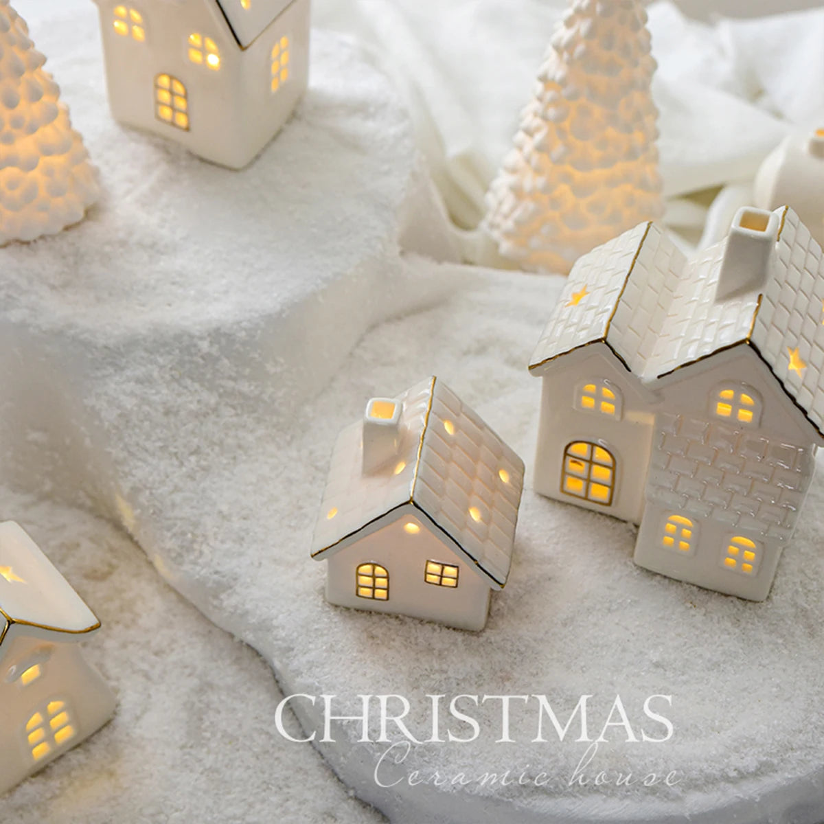 A set of ceramic Christmas houses with lights, designed to create a mini Christmas village scene, placed on a surface with snow and decorative lights.