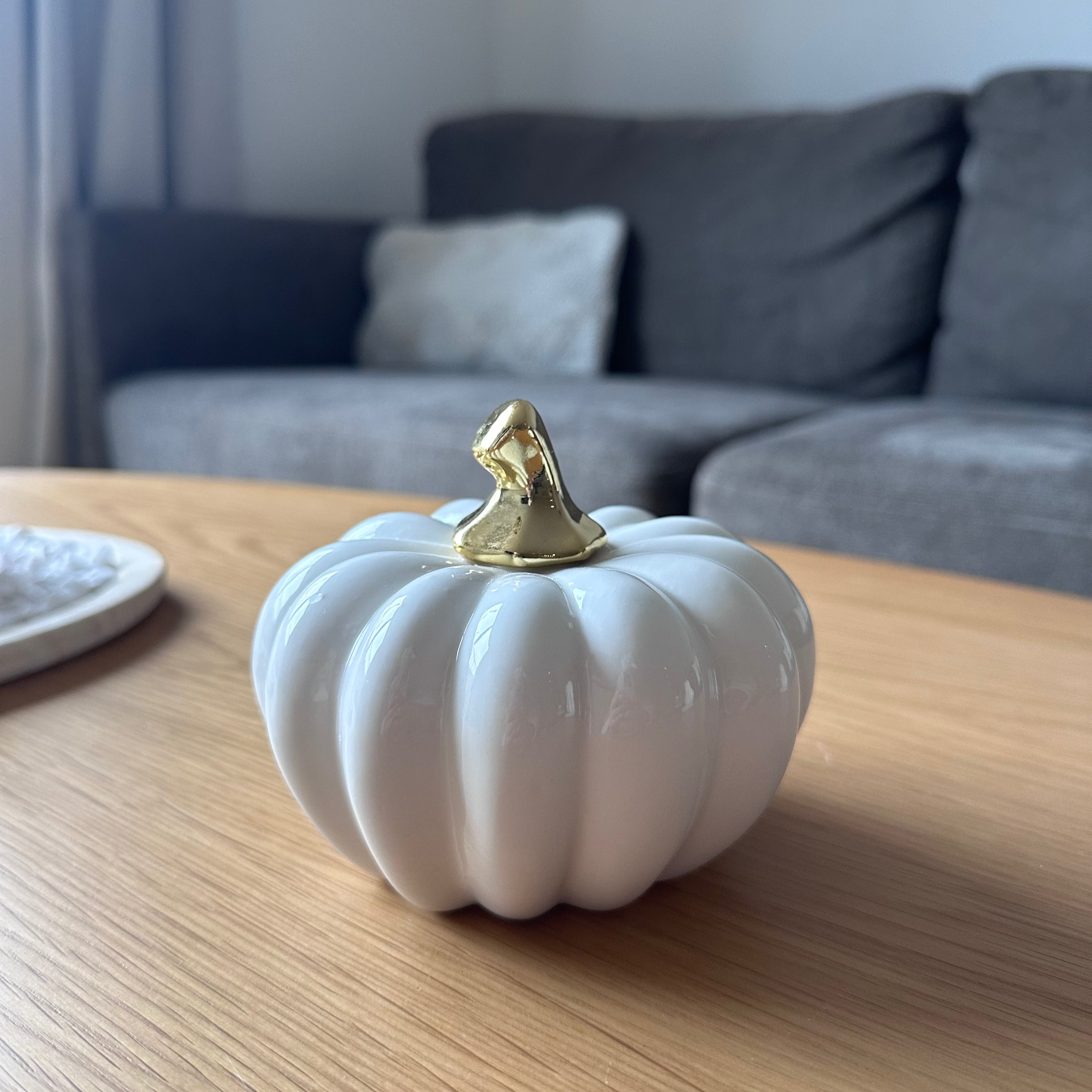 White ceramic pumpkin with a gold top on a wooden table, blurred sofa in the background