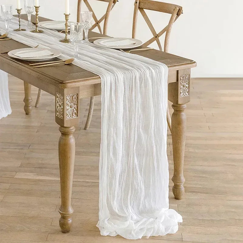 A white cheesecloth table runner with a rustic design, laid out on a wooden dining table with a decorative edge and floral pattern on the table.