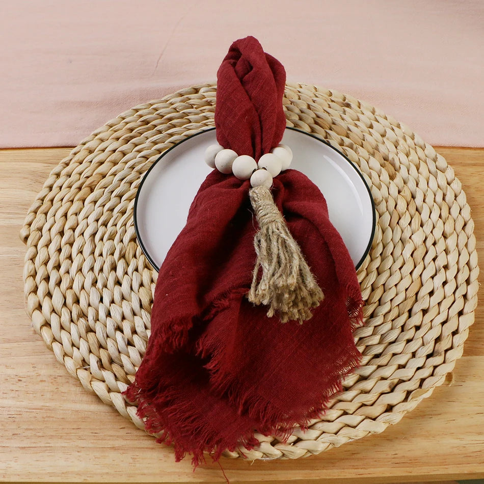 A burgundy cotton napkin is draped over a round woven basket, with a white plate and a beige decorative ball on top of the basket, set against a beige background.
