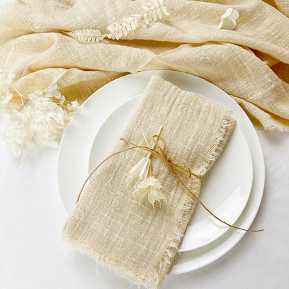 A beige cotton napkin is placed on a white plate, tied with a string to a decorative branch, with a neutral-toned textile in the background.