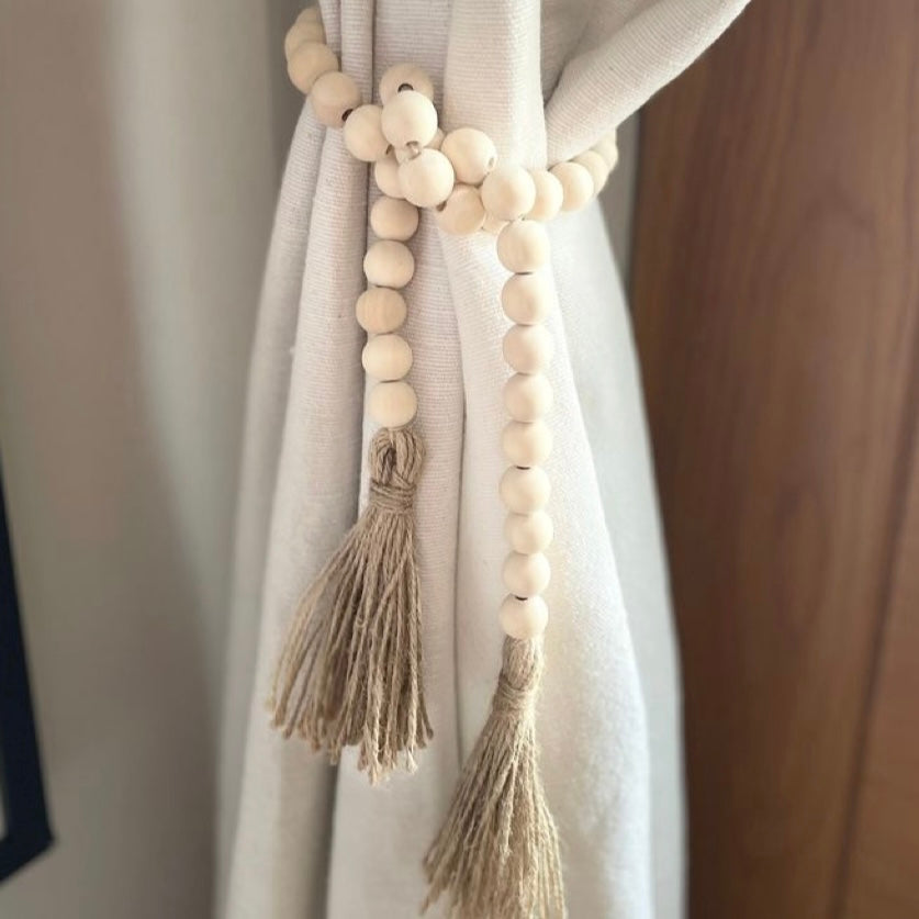 Wooden farmhouse beads decor, with a tassel tied around some beige minimalist curtains. 