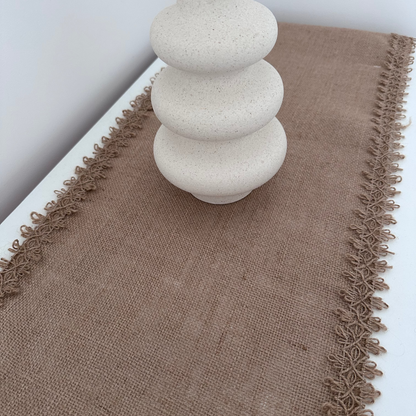 Brown textured, jute table runner with decorative edge on a white surface, featuring a white candle ceramic wavy vase.
