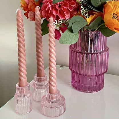 Pink twisted candles in pink glass holders with a pink vase containing flowers on a white surface.