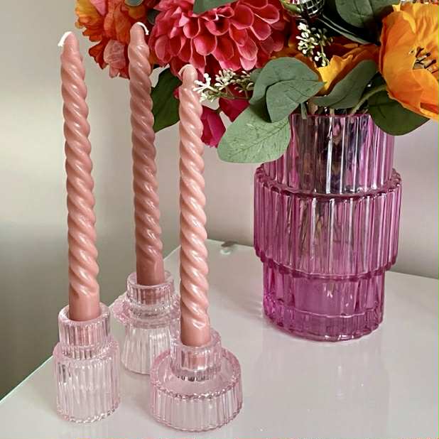 Pink twisted candles in pink glass holders with a pink vase containing flowers on a white surface.