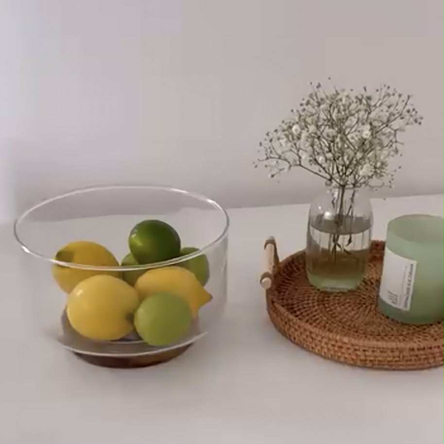 Clear glass bowl with lemons and limes on a white surface next to a woven tray with a plant and candle.