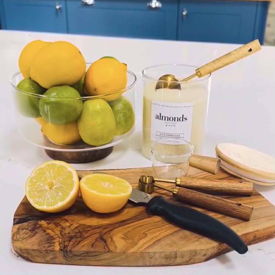 Fruit bowl with lemons and limes, a glass of almond milk, and kitchen tools on a white surface.