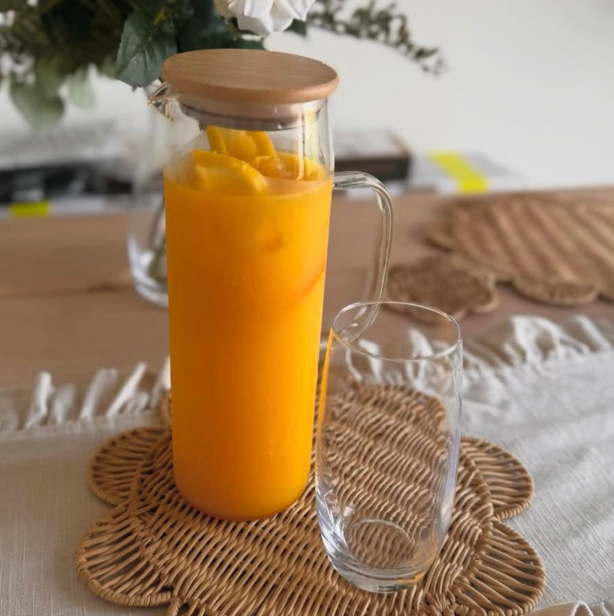 A clear glass water jug with a bamboo lid, filled with orange juice on a textured surface