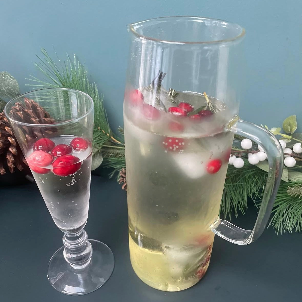 A clear glass water jug, filled with water and floating cherries. A pine cone wreath lays in the background. 