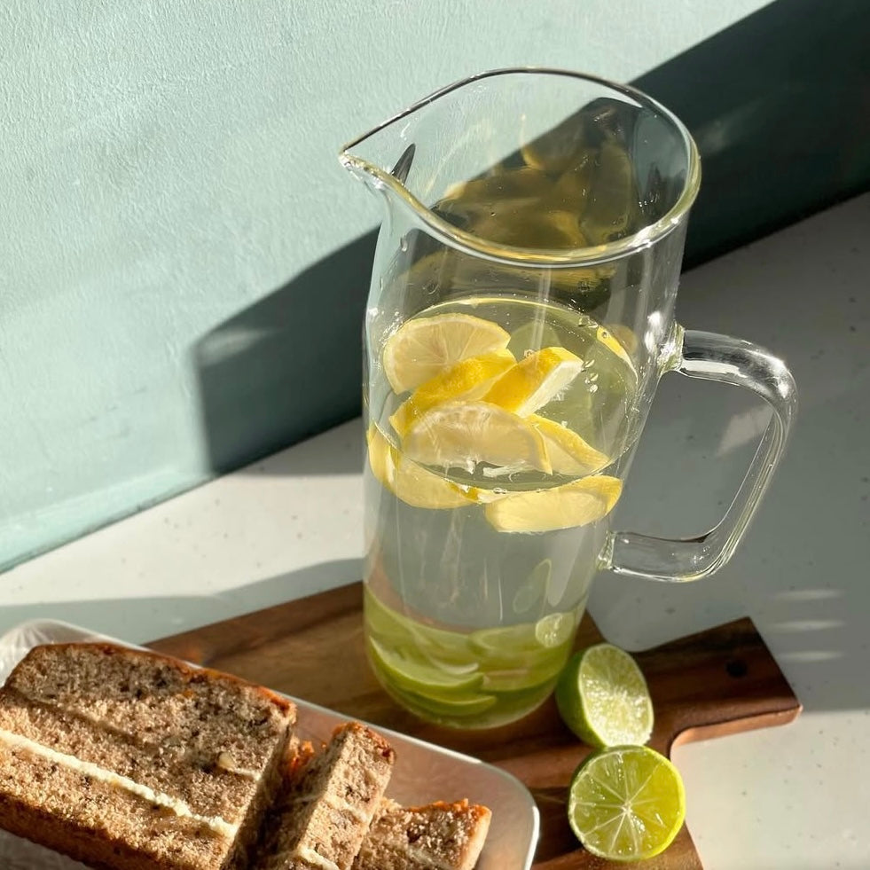 A clear glass water jug with a bamboo lid, filled with water and lemon slices, placed on a wooden board next to a slice of lime and some bread.