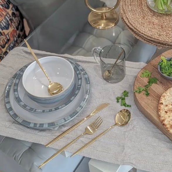 Dining table setting with gray plates, gold cutlery, and a glass on a light-colored tablecloth.