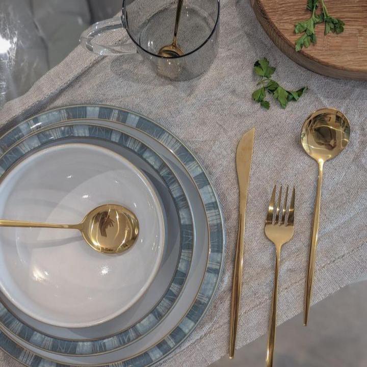 Elegant table setting with white & blue plates, gold cutlery, and a pink napkin on a textured table runner.