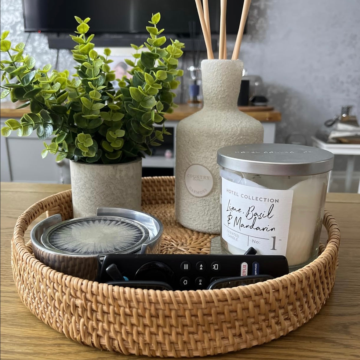 Handwoven, circular rattan tray styled with decorative items including a candle, diffuser, and plant.