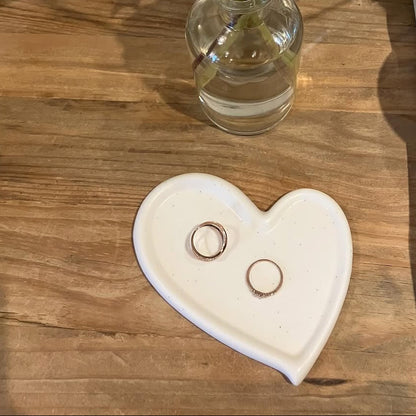 Heart-shaped dish with rings on a wooden surface next to a vase with flowers.