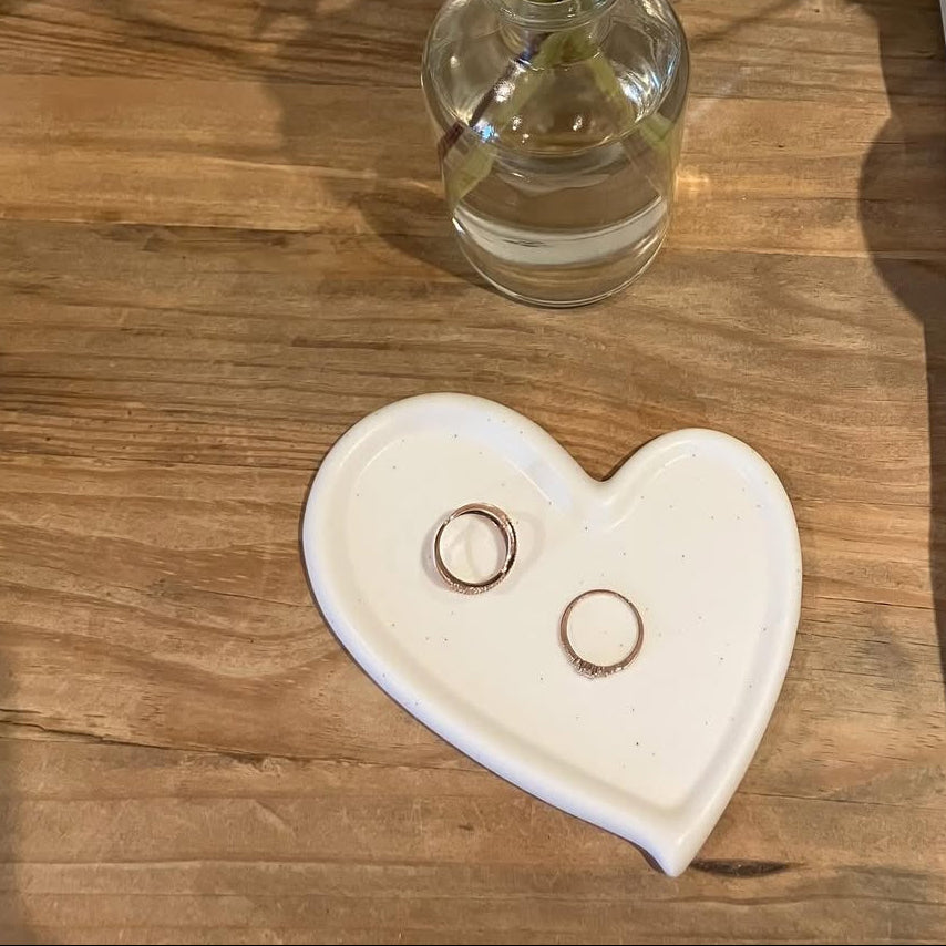 Heart-shaped dish with rings on a wooden surface next to a vase with flowers.