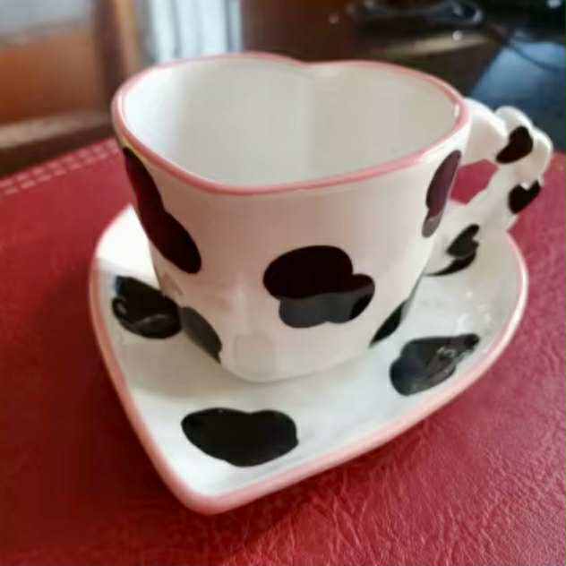 Heart-shaped ceramic cup and saucer with hand-painted, black cow print patterns on a red surface