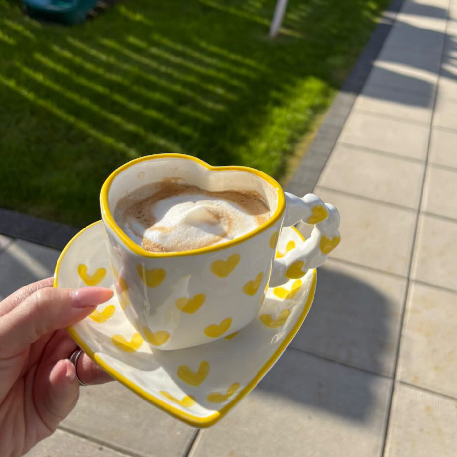 Ceramic heart-shaped mug with yellow , hand-painted heart patterns filled with a creamy beverage, held outdoors.