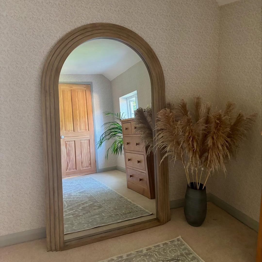 Wooden arched mirror on a wall with a vase of pampas grass to the right.