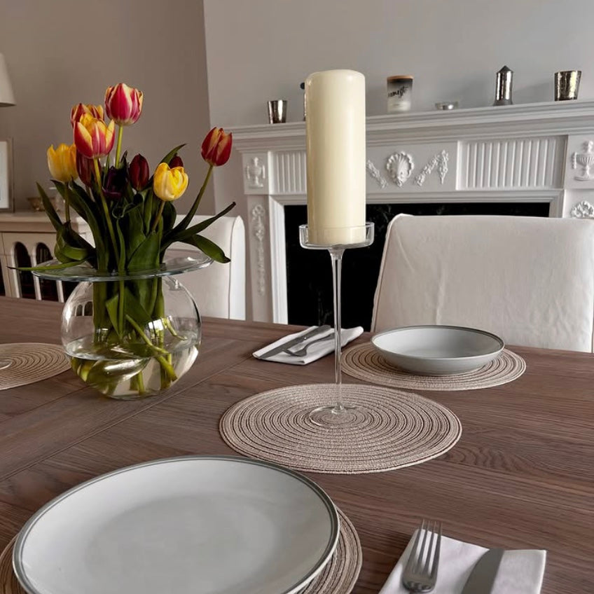 Dining table setting with plates, linen, woven placemats, and a vase of tulips in a room with a fireplace.