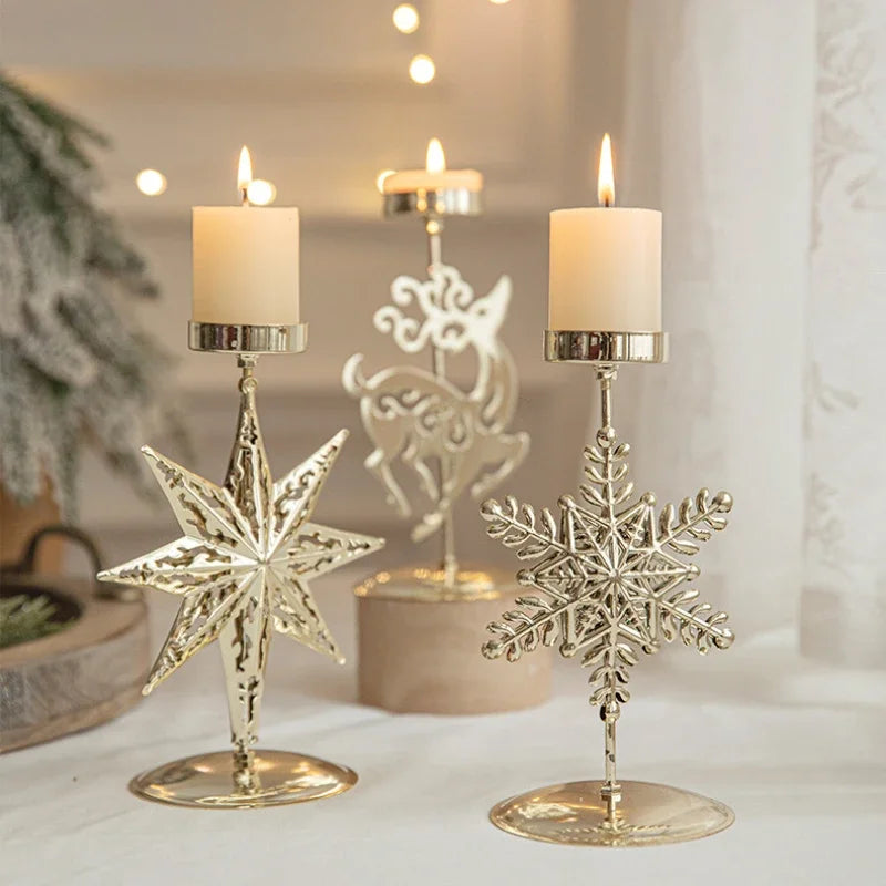 Two metal candle holders with gold engravings, one holding a lit candle, the other with a snowflake design, placed on a white surface with a blurred Christmas tree in the background.