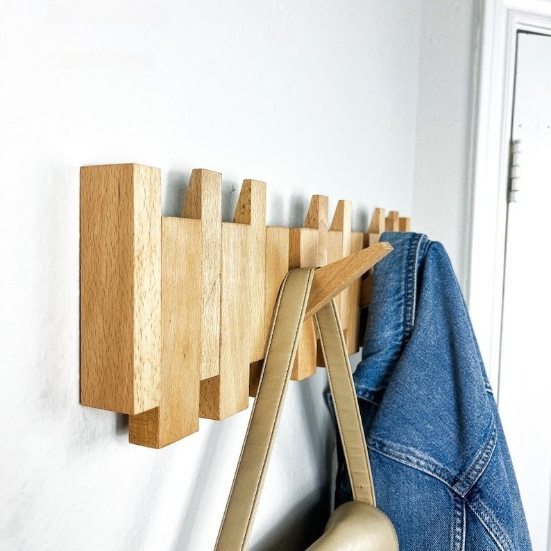 Wooden wall-mounted coat rack with hooks holding a denim jacket and a beige bag against a white wall.